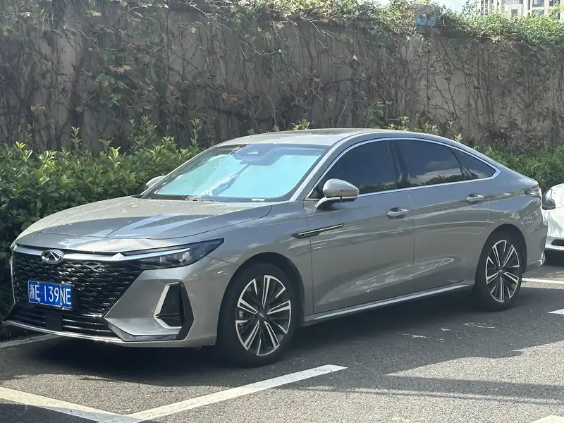 Chery Arrizo 8 סדאן אפור, מבט מהצד ומלפנים, בחניון ליד קיר ירוק