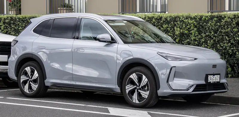Geely EX5 אפור בהיר, מבט מהצד ומלפנים, בחניון ליד גדר חיה